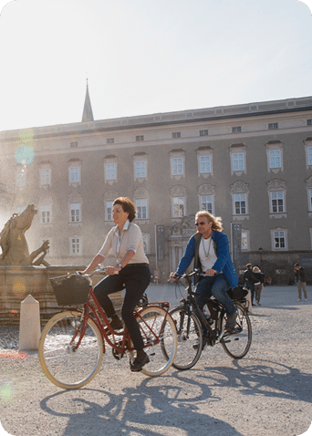 Zwei Frauen fahren mit Fahrrädern durch die Salzburger Altstadt bei Sonnenschein.