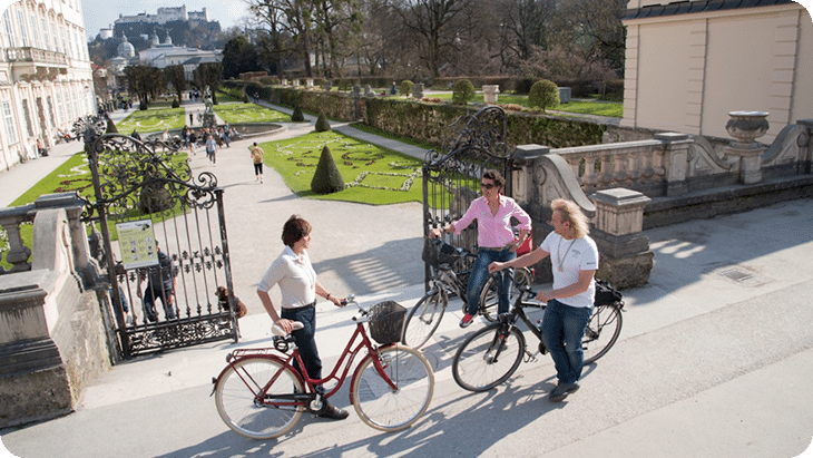 Drei Personen mit Fahrrädern unterhalten sich im Mirabellgarten mit Blick auf die Festung Hohensalzburg.